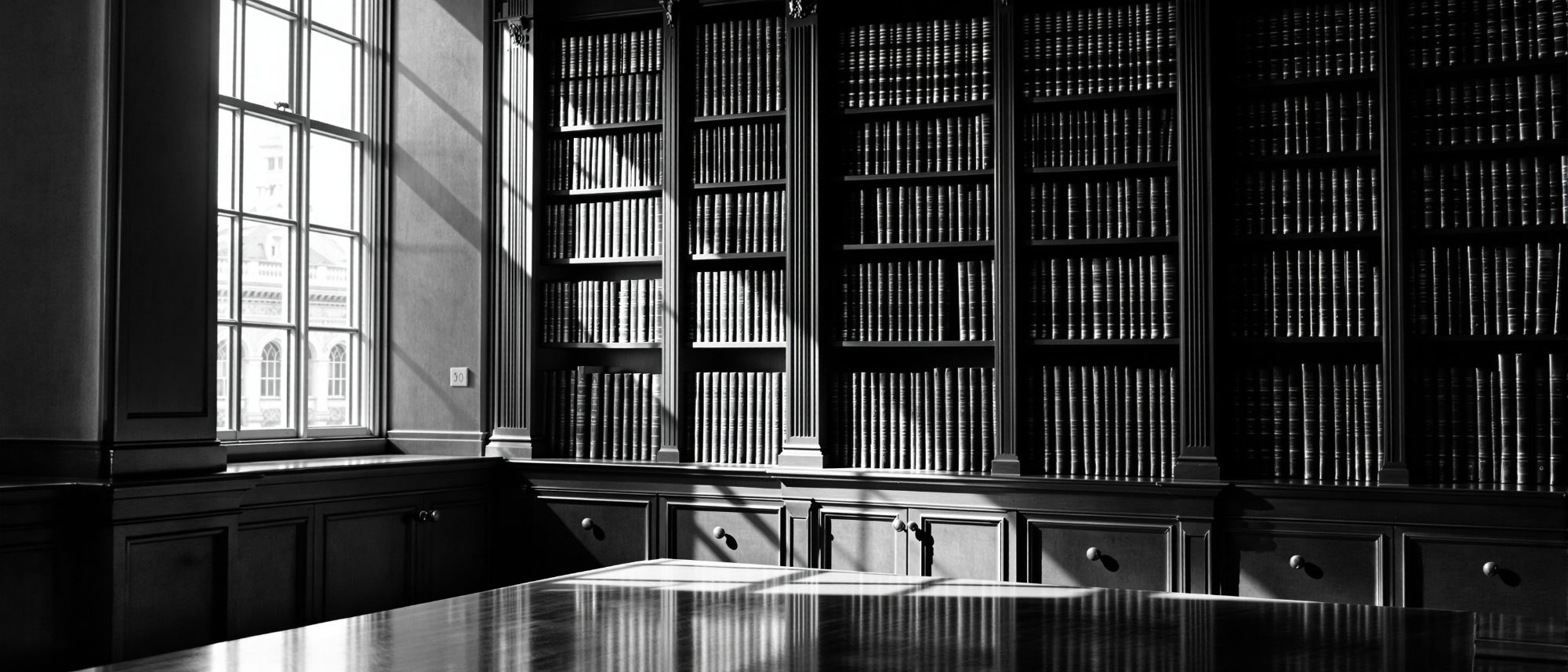 Biblioteca jurídica con estanterías de volúmenes encuadernados y luz lateral
        entrando por un ventanal alto.